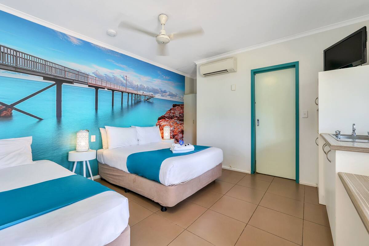 Habitación de hotel con dos camas, un mural de un muelle, ventilador de techo, aire acondicionado, televisor y baño con lavamanos y espejo.