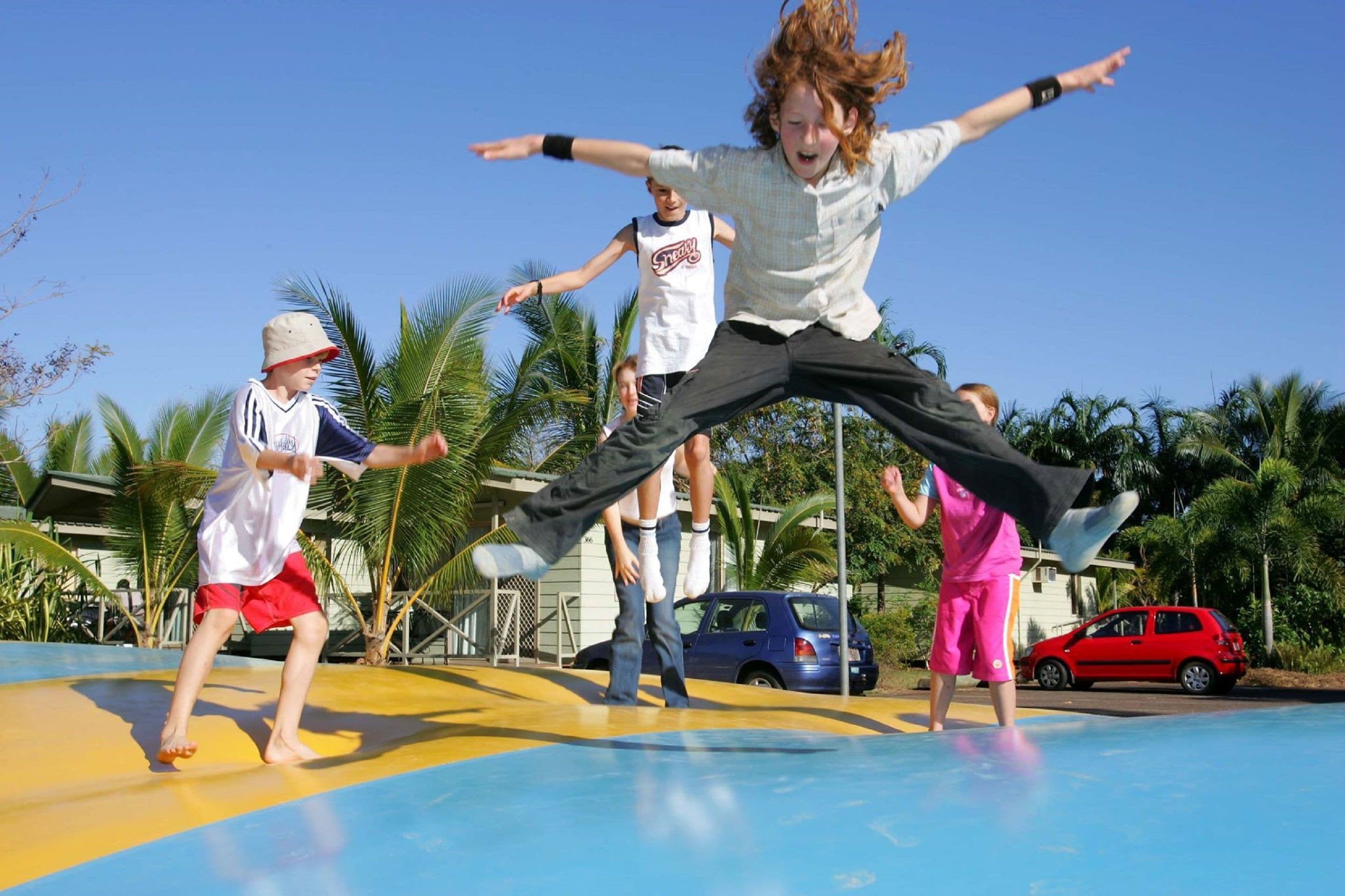 A group of people jumping on a trampoline outdoors with palm trees and a car in the background.