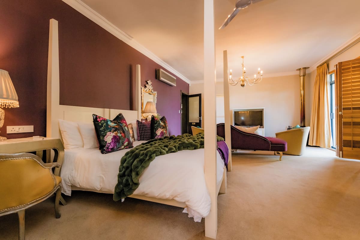A bedroom with a four-poster bed, a desk with a lamp, a chair, a television, and a window with blinds.