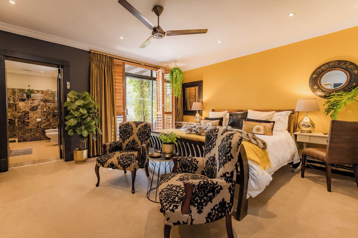 A bedroom with a bed, two armchairs, a side table, a mirror, and a ceiling fan.