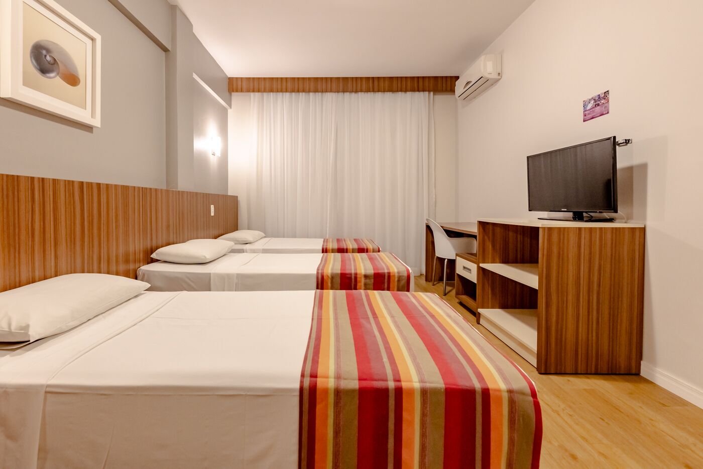 Quarto de hotel com três camas, uma escrivaninha, televisão e janela com cortinas.