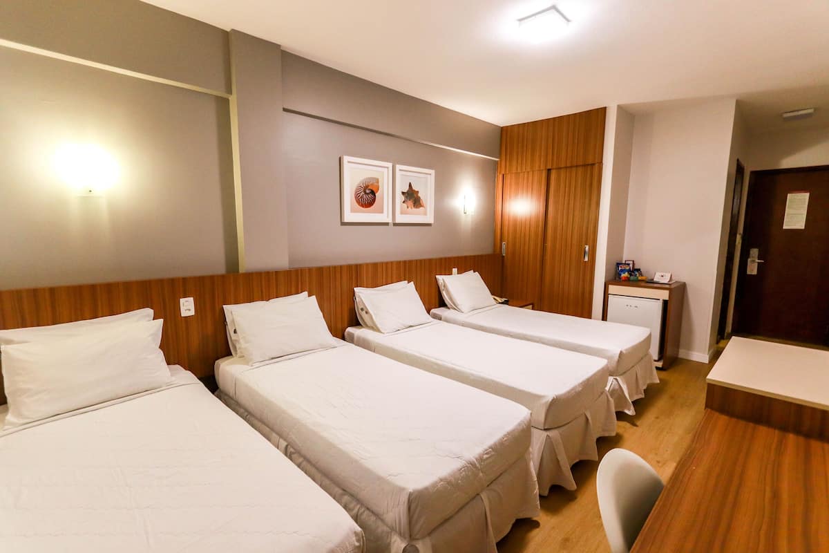 Quarto de hotel com três camas de solteiro, um guarda-roupa de madeira, uma escrivaninha com cadeira e duas fotos emolduradas na parede.