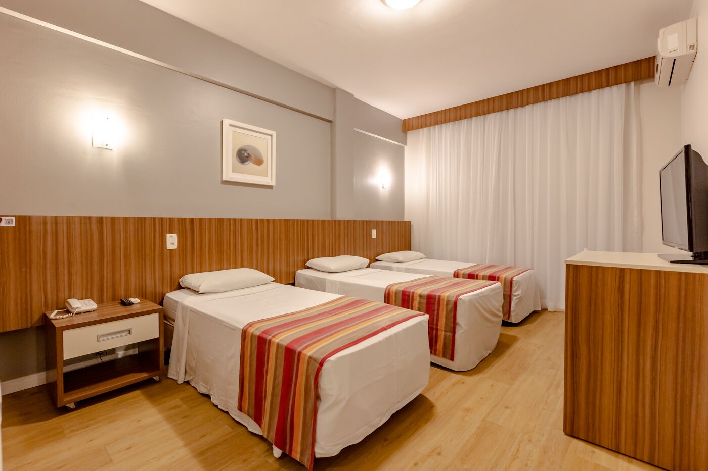 A hotel room with three single beds, a wooden headboard, a bedside table with a phone, a TV on a wooden stand, and a large window with curtains.