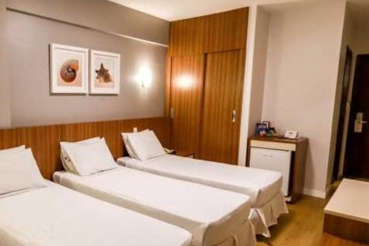 Quarto de hotel com duas camas de solteiro, um guarda-roupa de madeira, uma pequena escrivaninha com telefone e duas imagens emolduradas na parede.