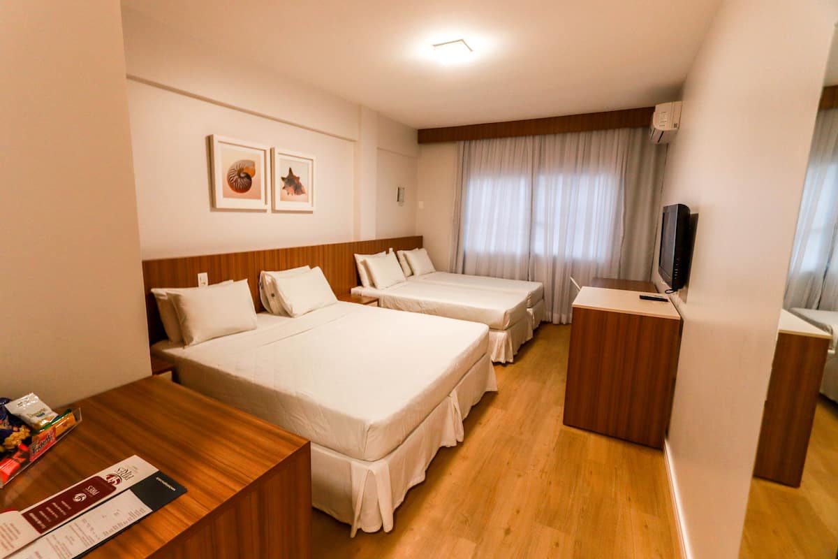 Quarto de hotel com duas camas, uma escrivaninha de madeira, televisão e uma janela com cortinas.