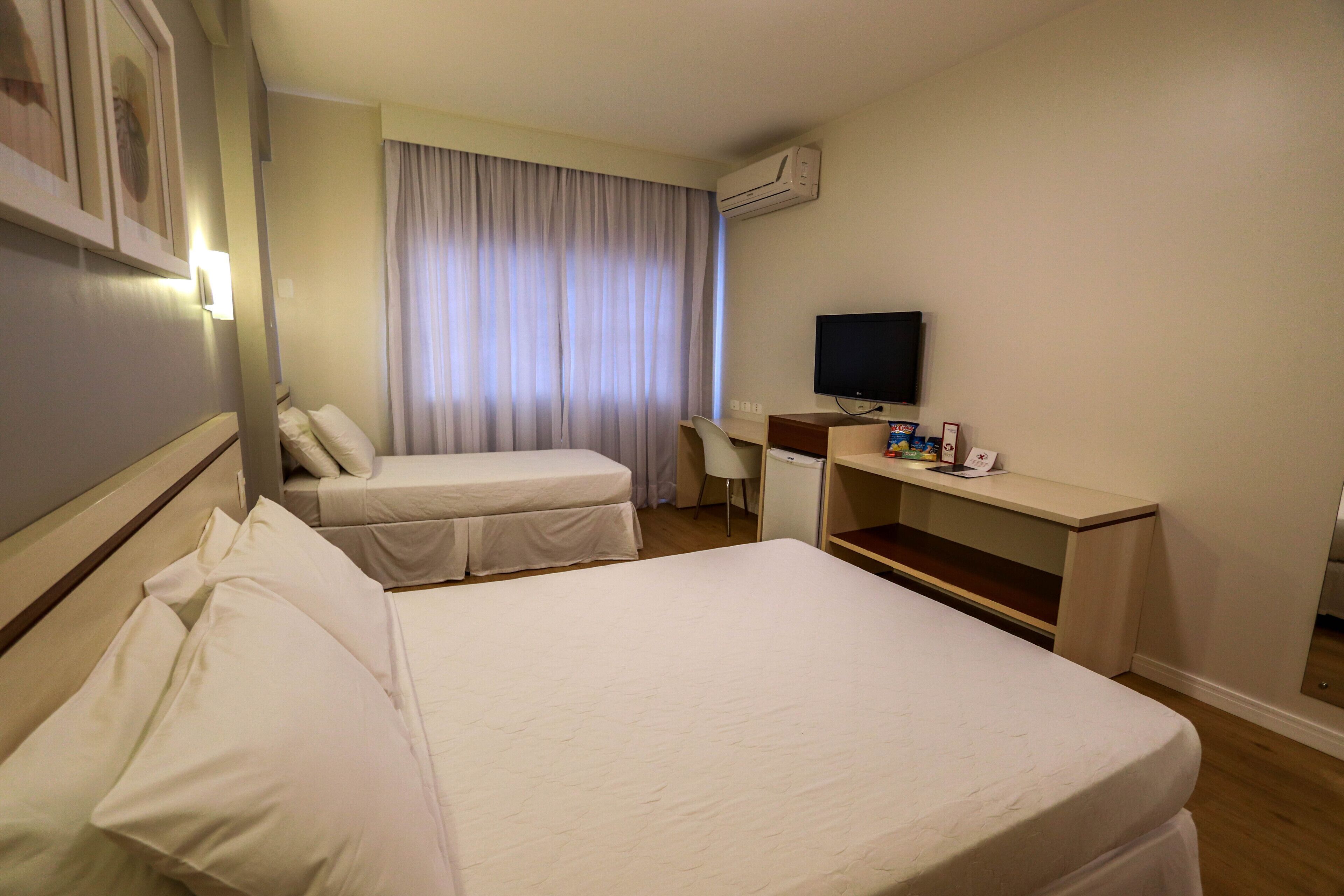 A hotel room with two beds, a desk, a television, and an air conditioning unit.