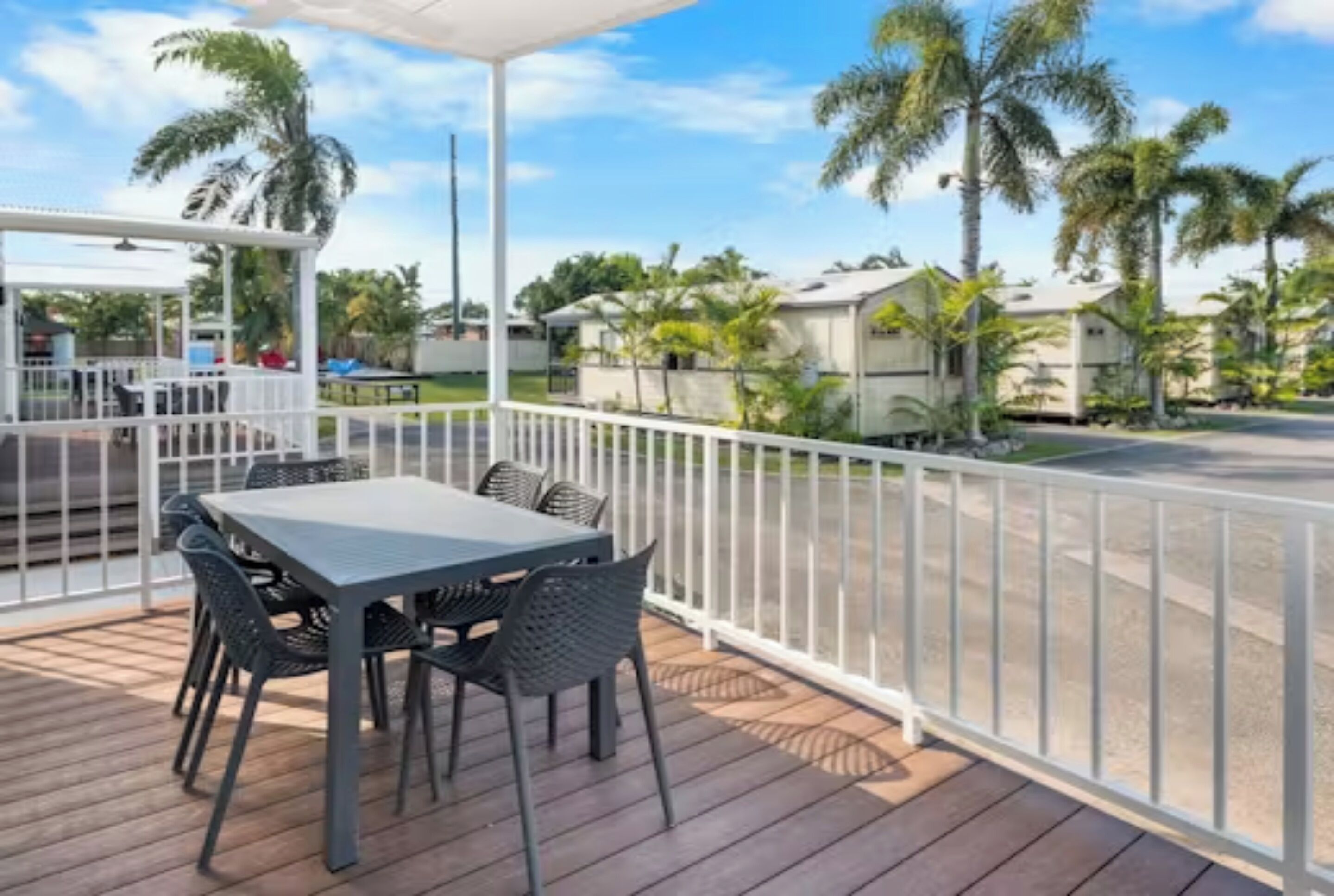 A covered outdoor area with a table and chairs, surrounded by a white railing, overlooking a street with houses and palm trees.