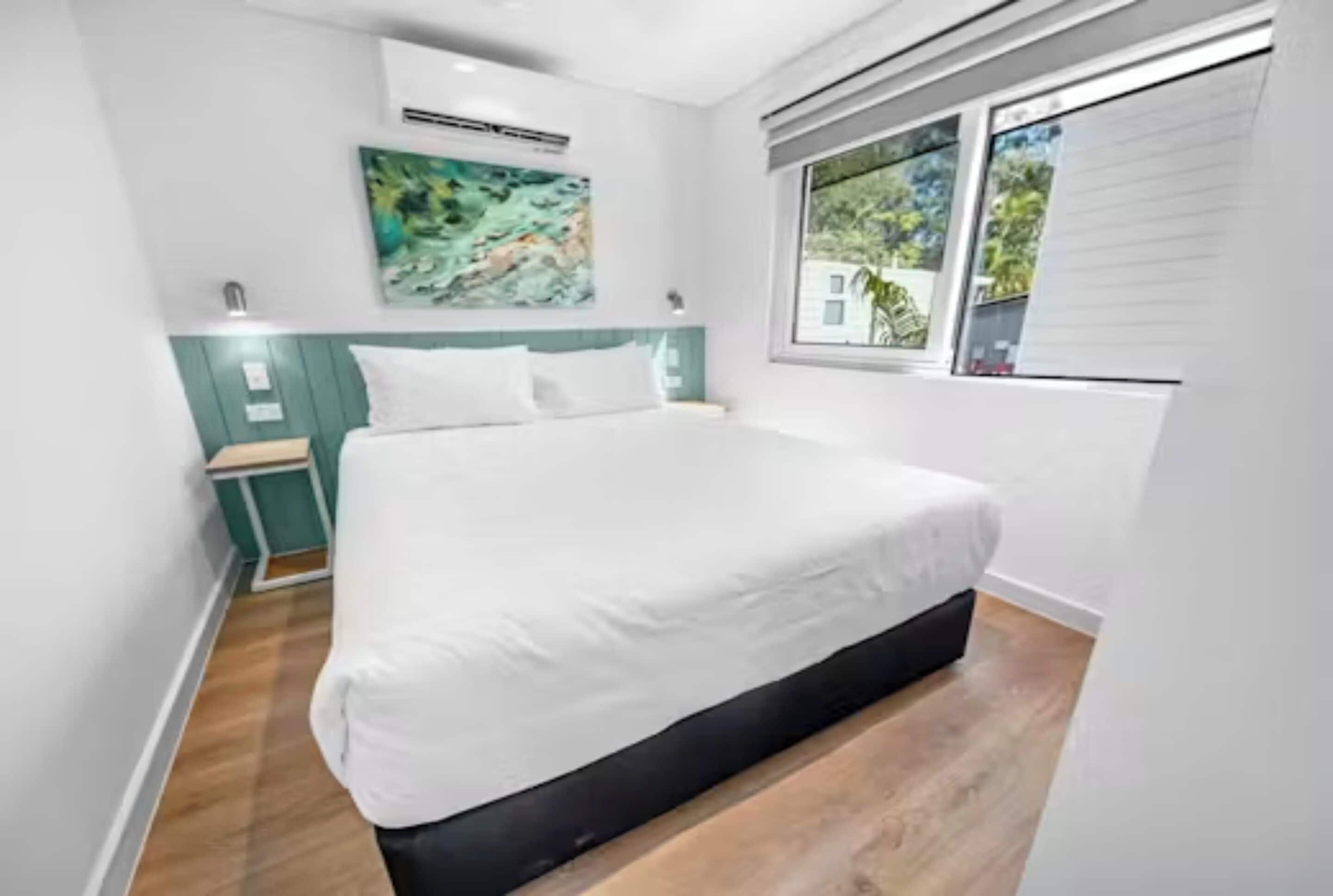 A modern bedroom with a bed, a window with blinds, a wall-mounted air conditioner, and a painting.