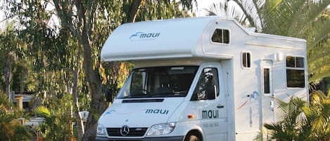 Un camping-car blanc de la marque « maui » est garé dans une zone herbeuse, bordée d’arbres, sous un ciel dégagé.