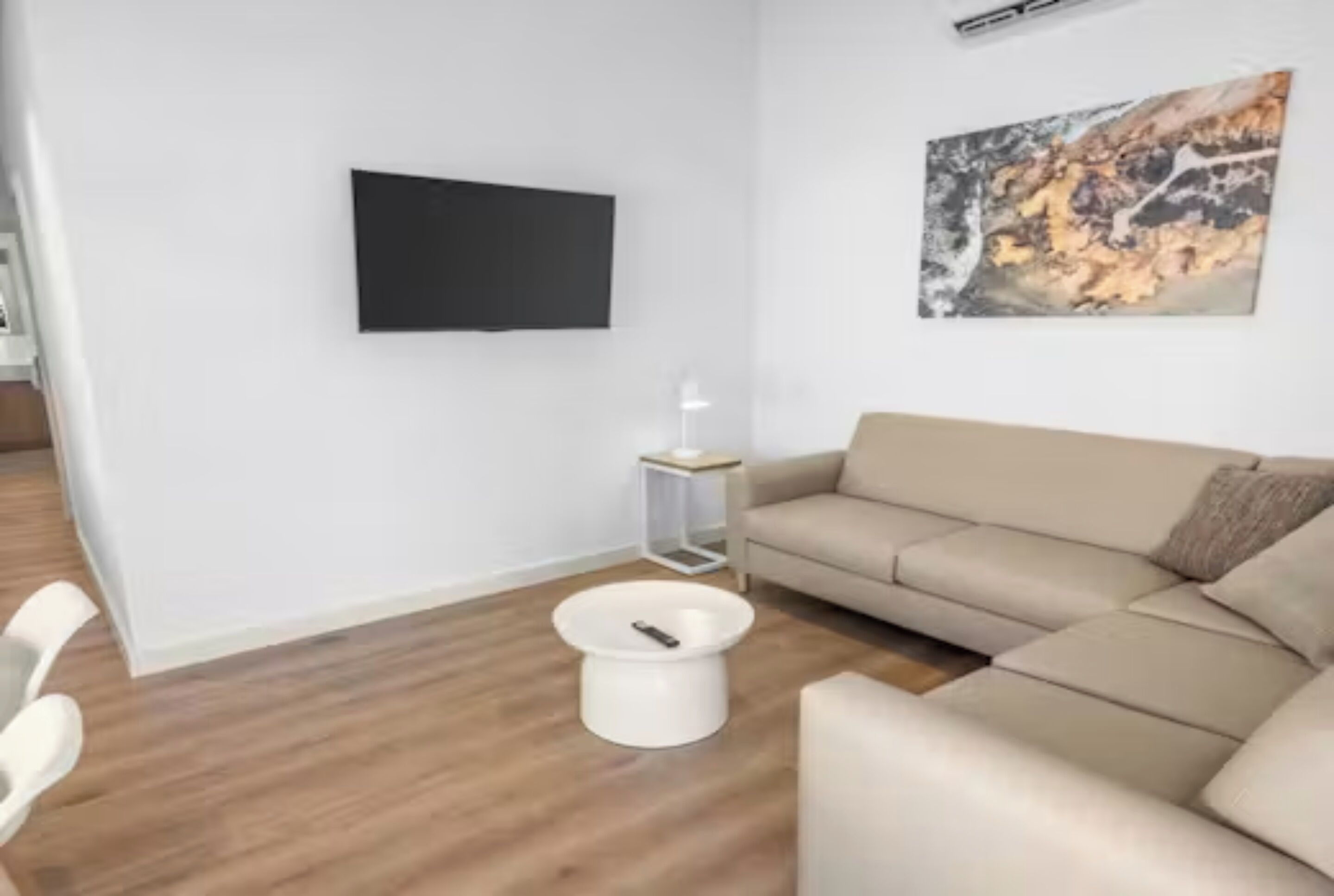 A modern living room with a beige sofa, a flat-screen TV mounted on the wall, and a small side table with a lamp.