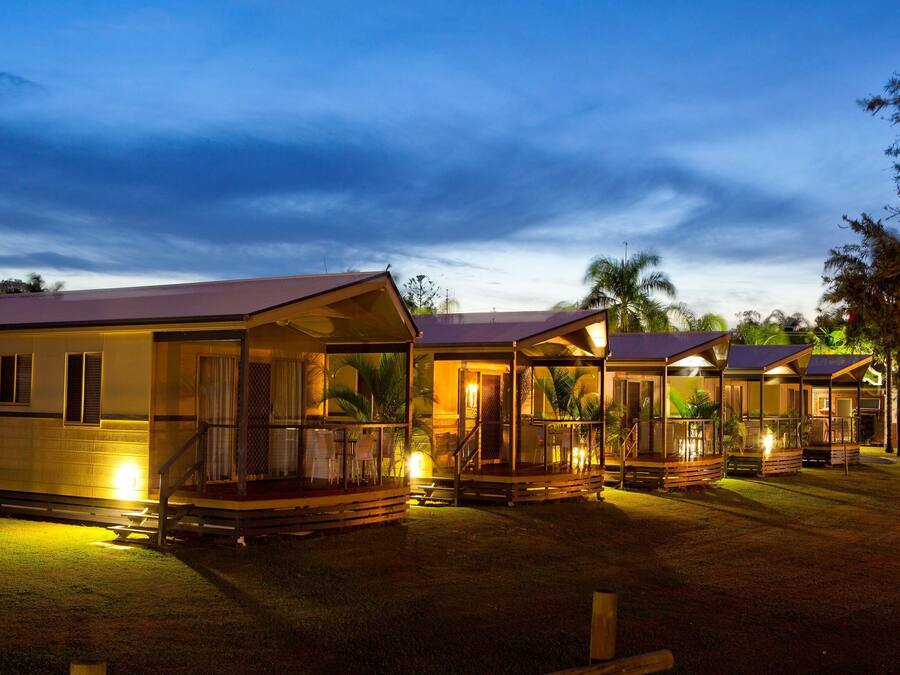 A row of cabins with glass fronts and wooden decks, set in a grassy area with palm trees in the background.