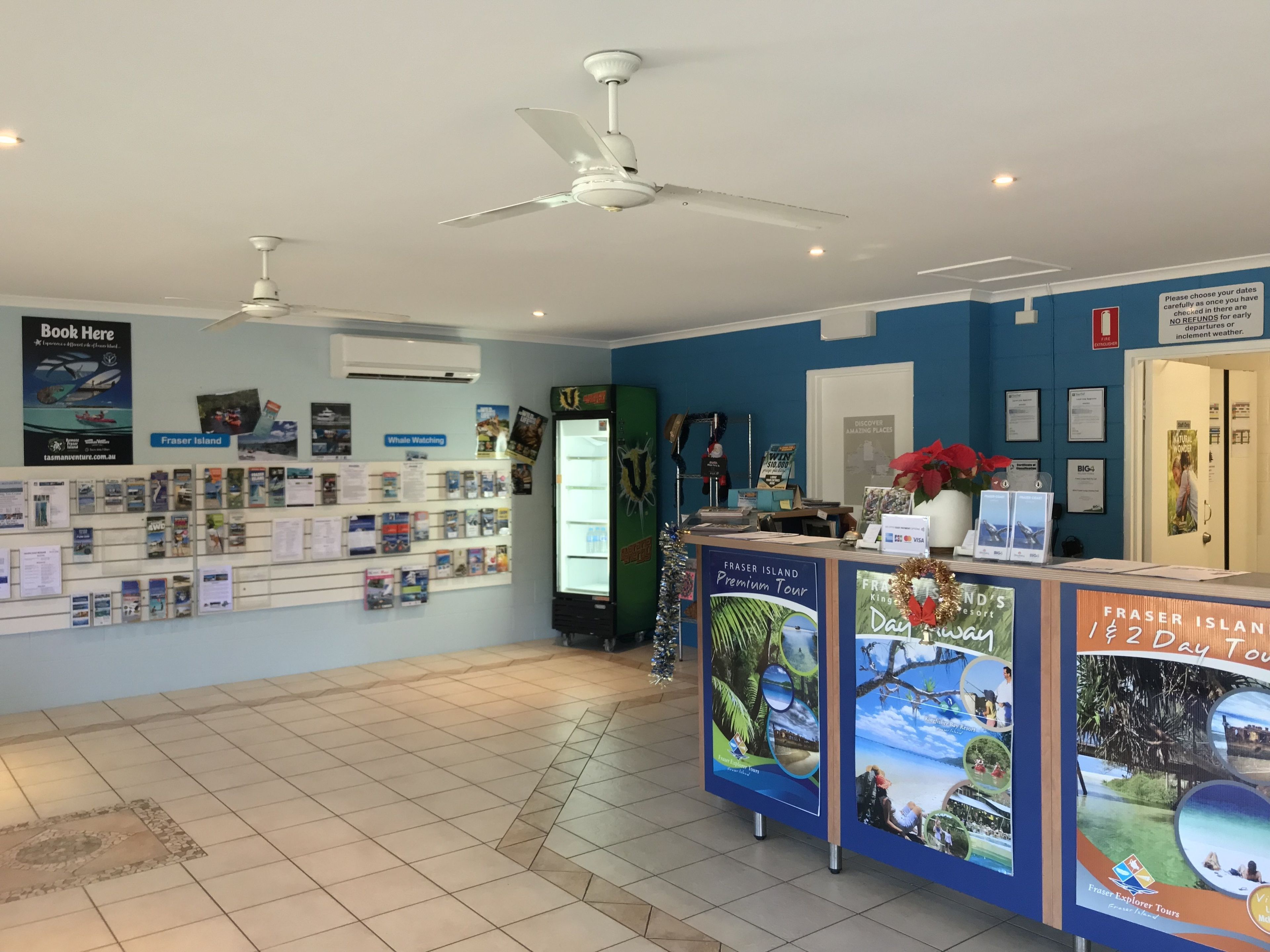 Un espace d’accueil avec un comptoir où sont exposées des brochures de voyage, un mur orné d’affiches de voyage et un ventilateur de plafond.