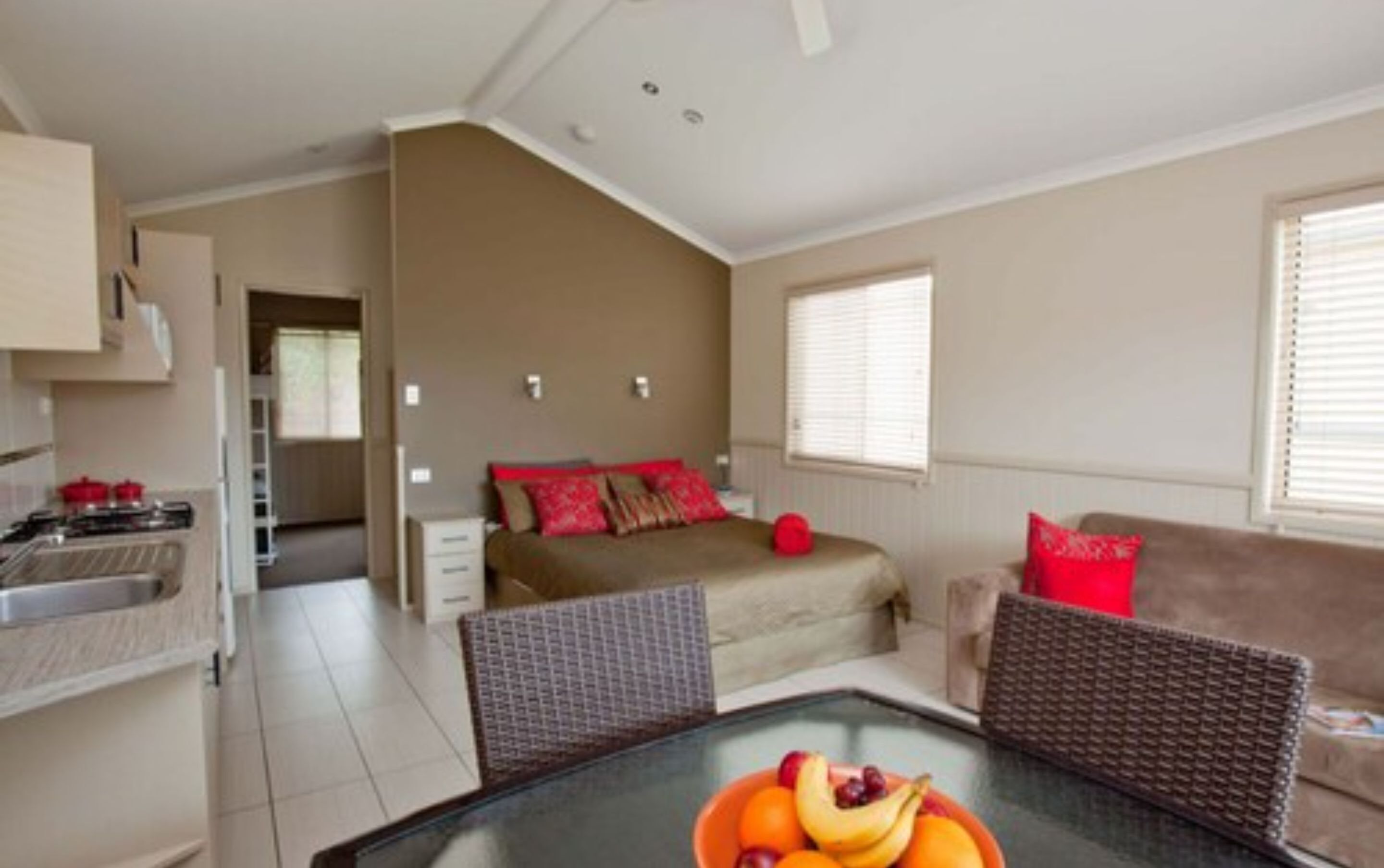 A compact living space with a kitchenette, a bed with red pillows, a dining table with fruit, and a sofa.