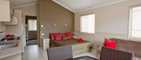 A compact living space with a kitchenette, a bed with red pillows, a dining table with fruit, and a sofa.