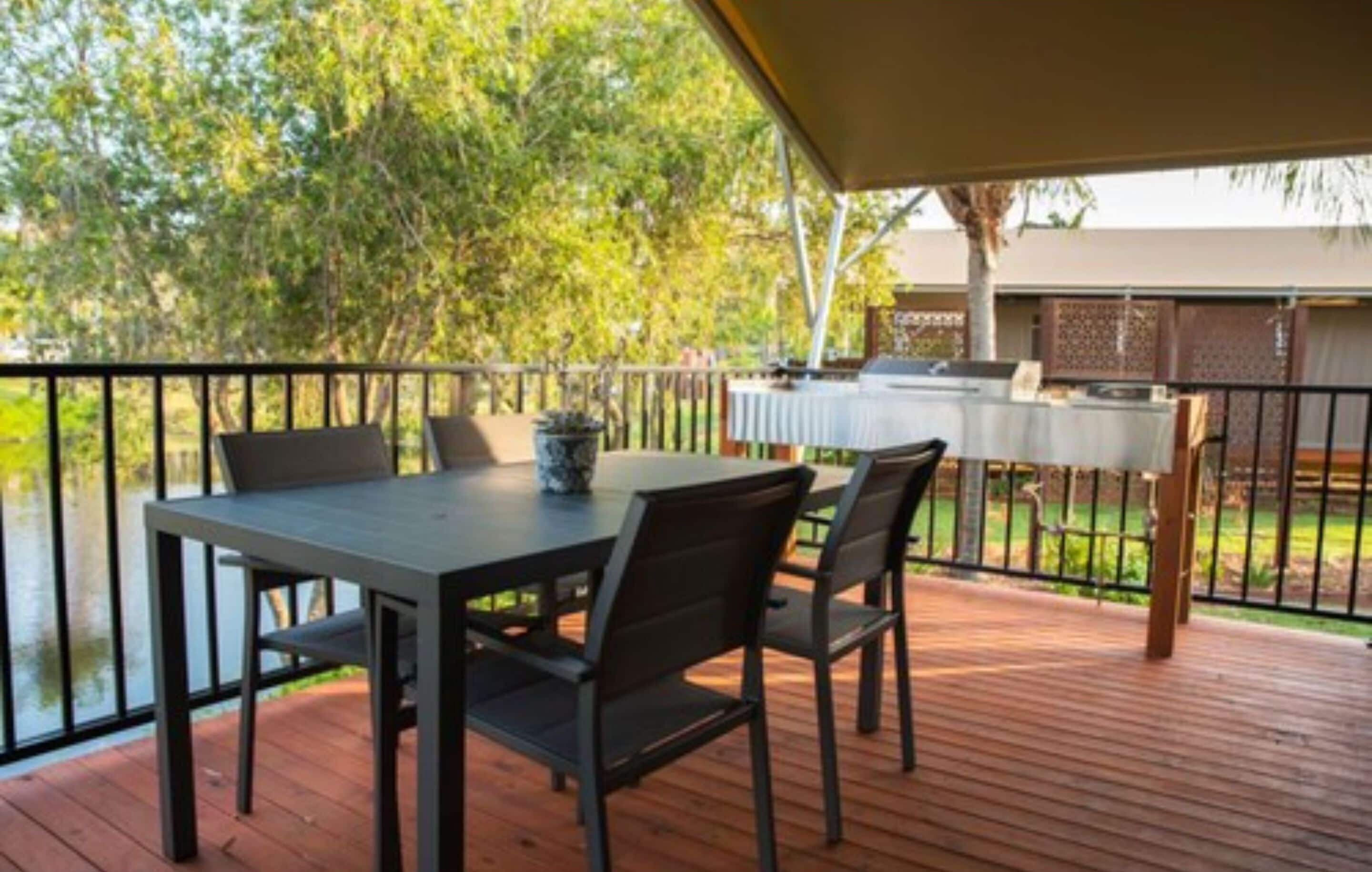 A covered outdoor patio with a table and chairs, overlooking a body of water and a fenced area with a grill.