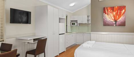 A hotel room with a bed, a desk, a chair, a TV, a painting, and a kitchenette.