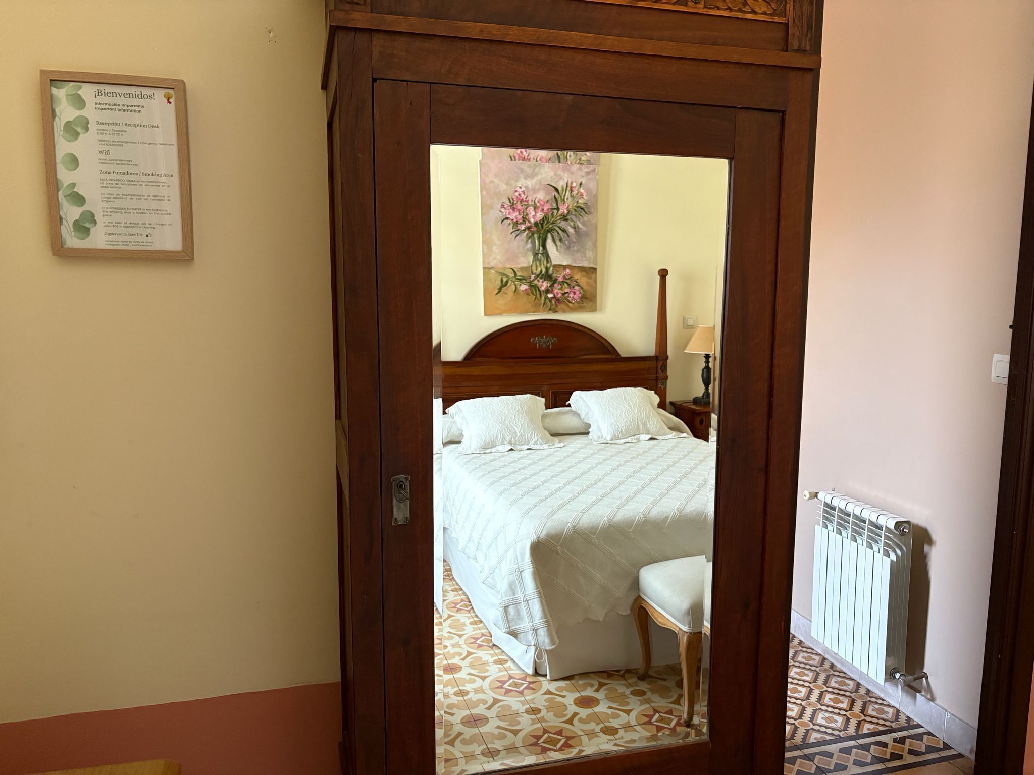 Habitación de hotel con puerta de madera, mostrador de recepción, un cuadro, cama con ropa blanca, una silla y un radiador.