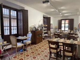 Uma área de refeições com várias mesas e cadeiras, um tapete estampado e grandes janelas com persianas de madeira.