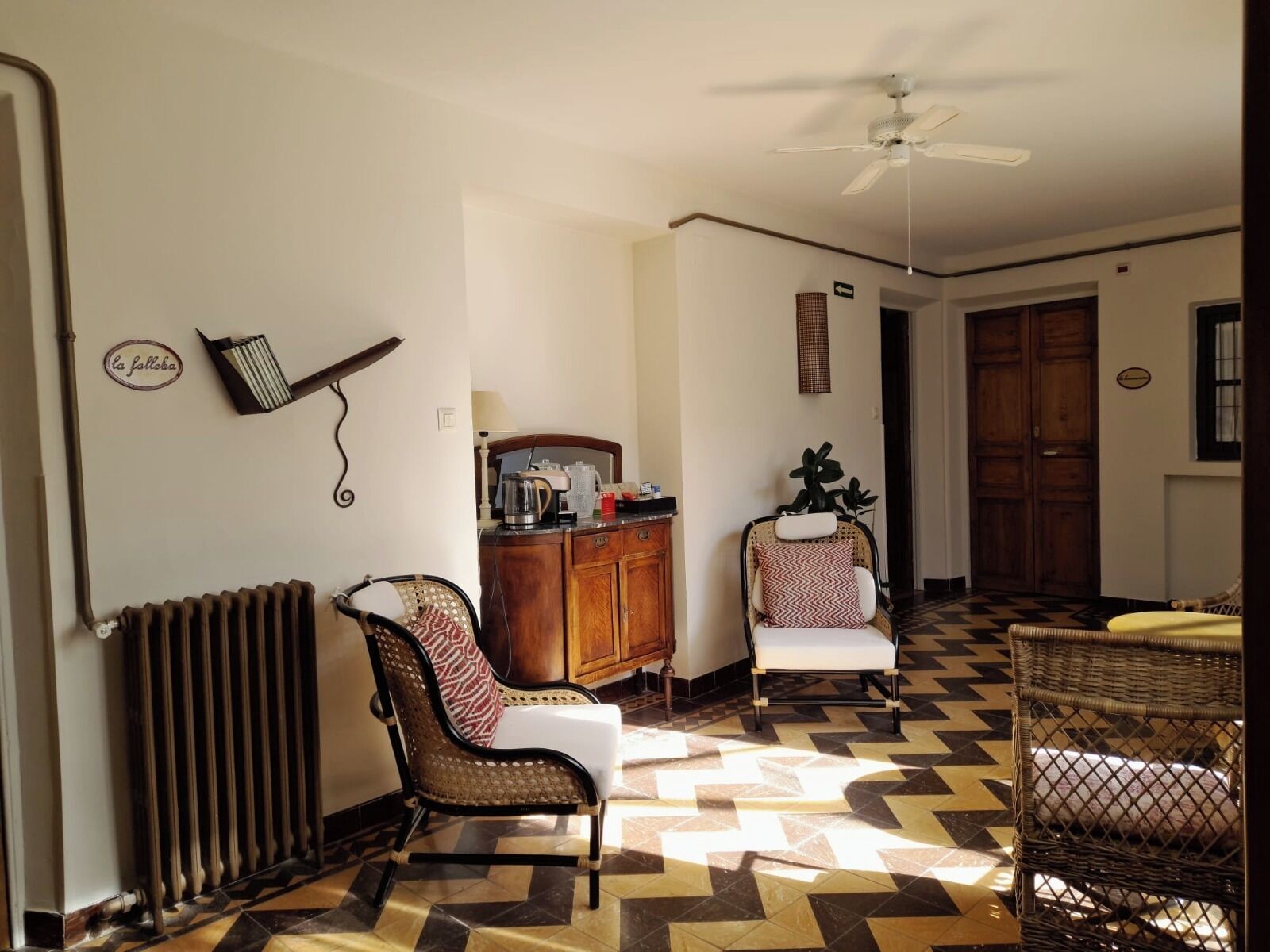 A room with a radiator, a ceiling fan, a cabinet with kitchen items, two chairs, and a patterned floor.