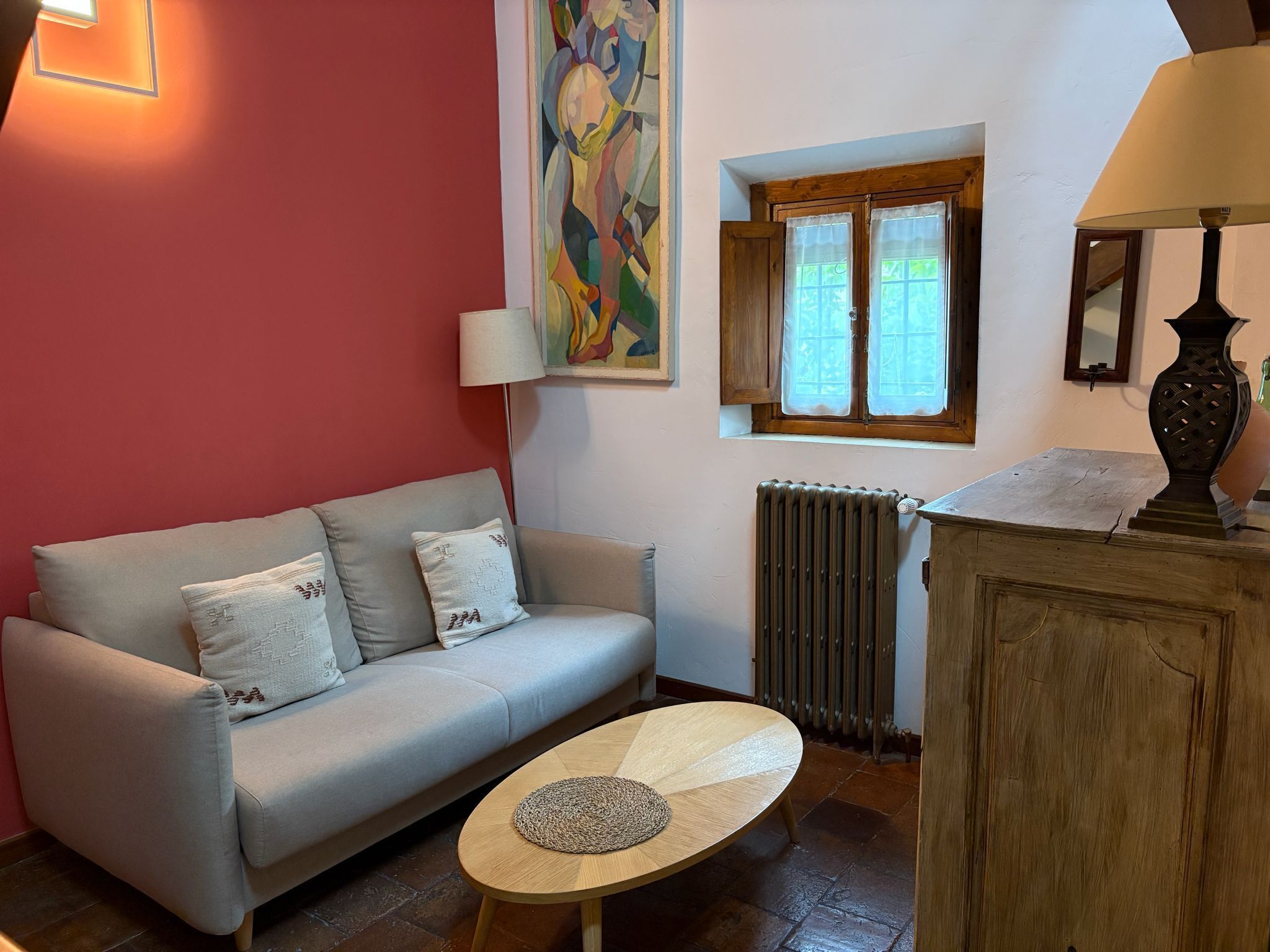 Un salon avec un canapé, une petite table ronde, un radiateur, une fenêtre avec des rideaux et un tableau accroché au mur.