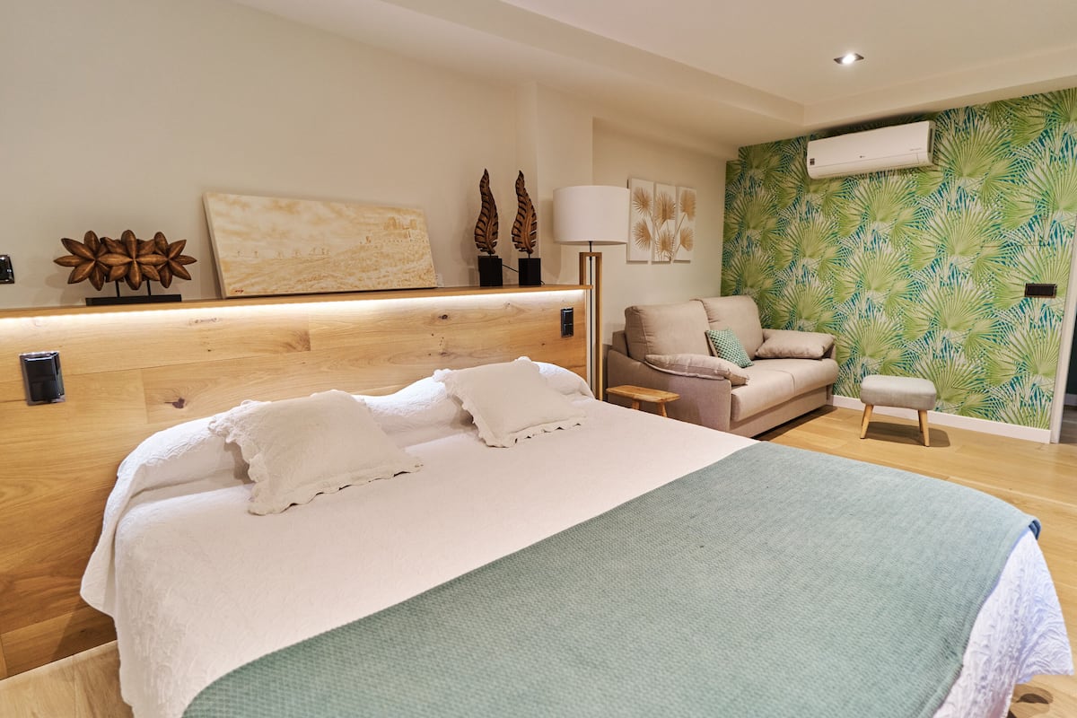 A modern bedroom with a large bed, a sofa, a small stool, and a wall with a tropical leaf pattern.