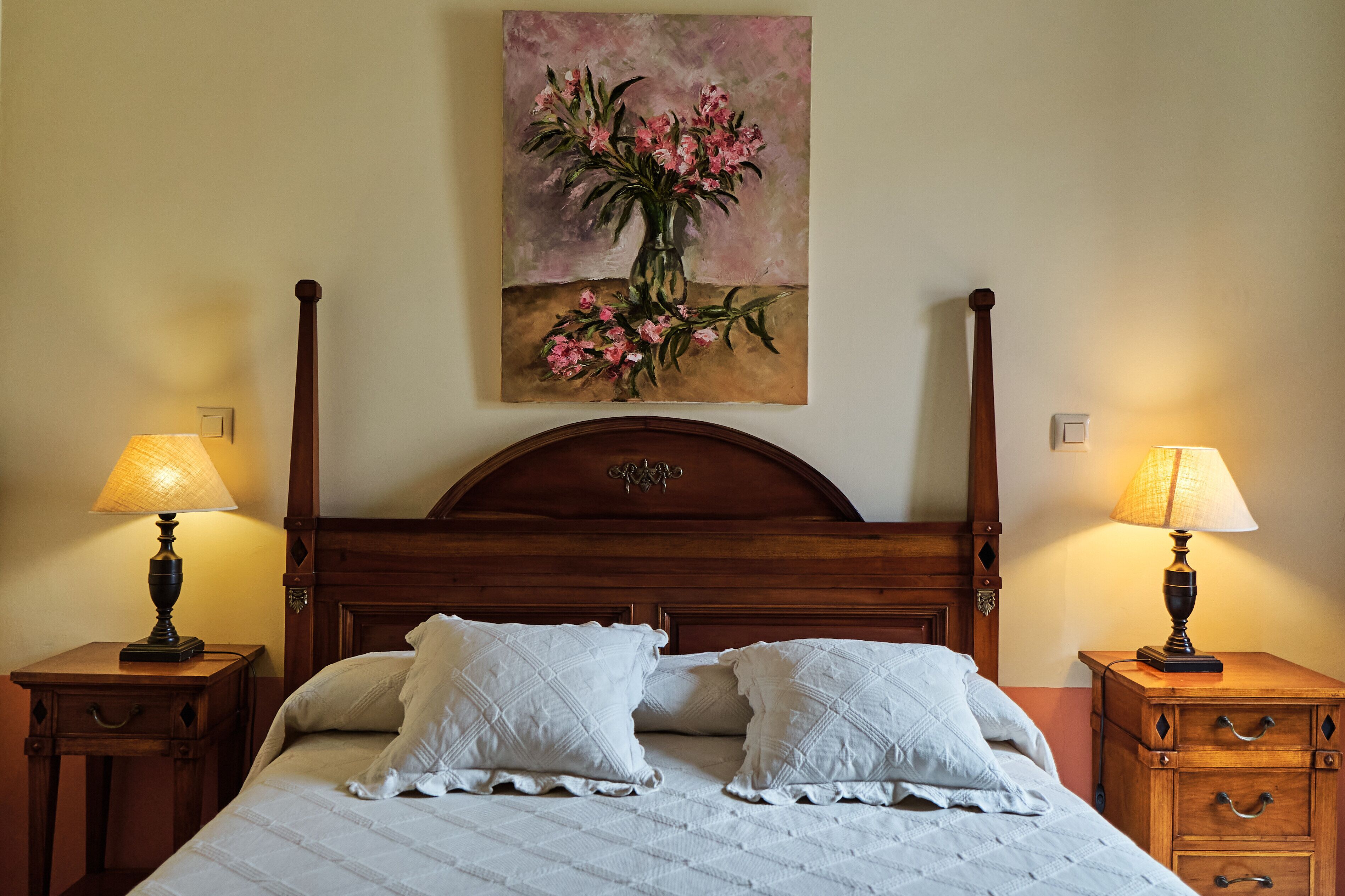 Un dormitorio con cabecera de madera, dos mesitas de noche con lámparas, un cuadro de flores en la pared y una cama con ropa blanca.