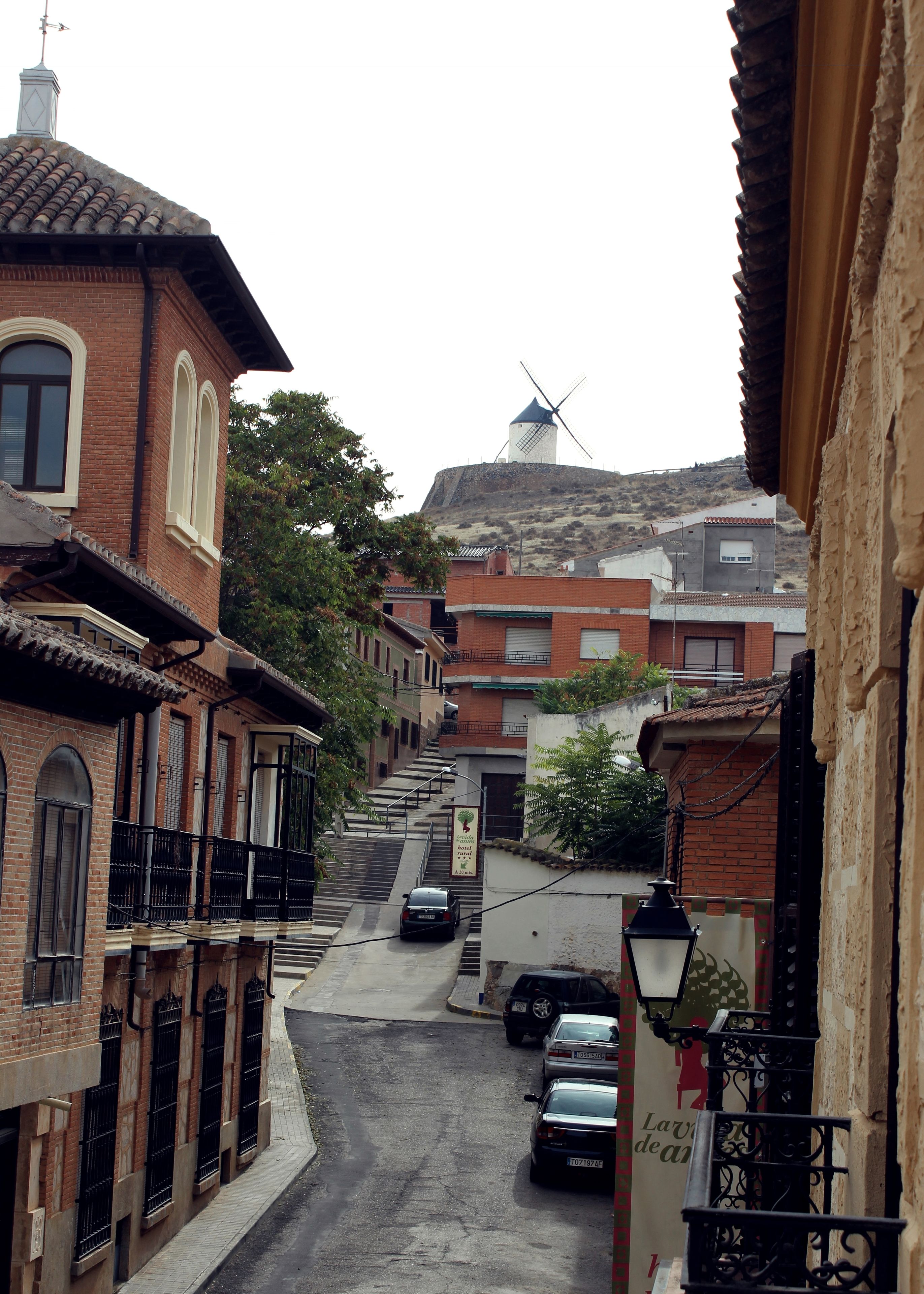 Une rue étroite bordée de bâtiments traditionnels, avec un moulin à vent et des voitures garées.