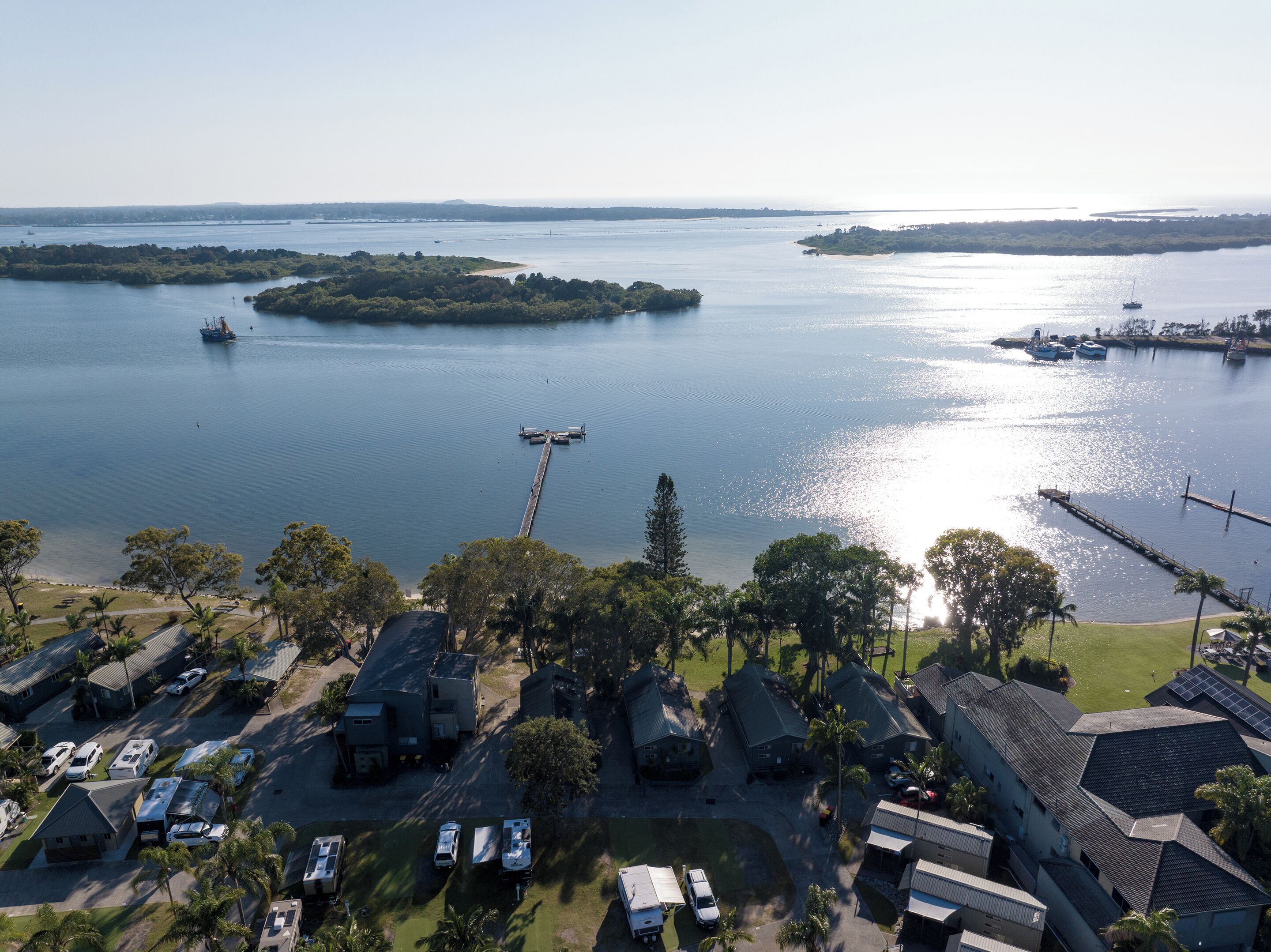 Eine Küstenlandschaft mit einer Marina, Booten und einer anlegenden Pier. Im Vordergrund befinden sich Wohngebäude und geparkte Fahrzeuge.