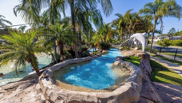 A swimming pool with a natural rock edge, surrounded by palm trees and a white dolphin statue. - NRMA Blue Dolphin Yamba Holiday Resort (Yamba)