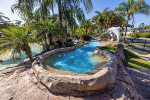 A swimming pool with a natural rock edge, surrounded by palm trees and a white dolphin statue.
