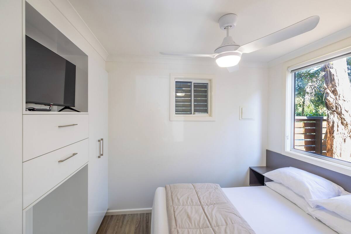 A bedroom with a bed, a ceiling fan, a window, and a TV on a cabinet.