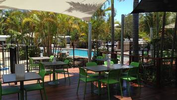 An outdoor patio area with wooden flooring, green plastic chairs, and tables. There is a pool in the background surrounded by palm trees.