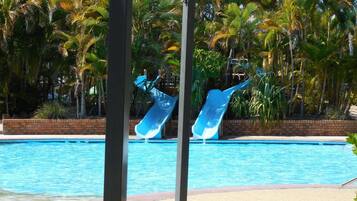 A swimming pool with two slides, surrounded by palm trees and a brick wall.