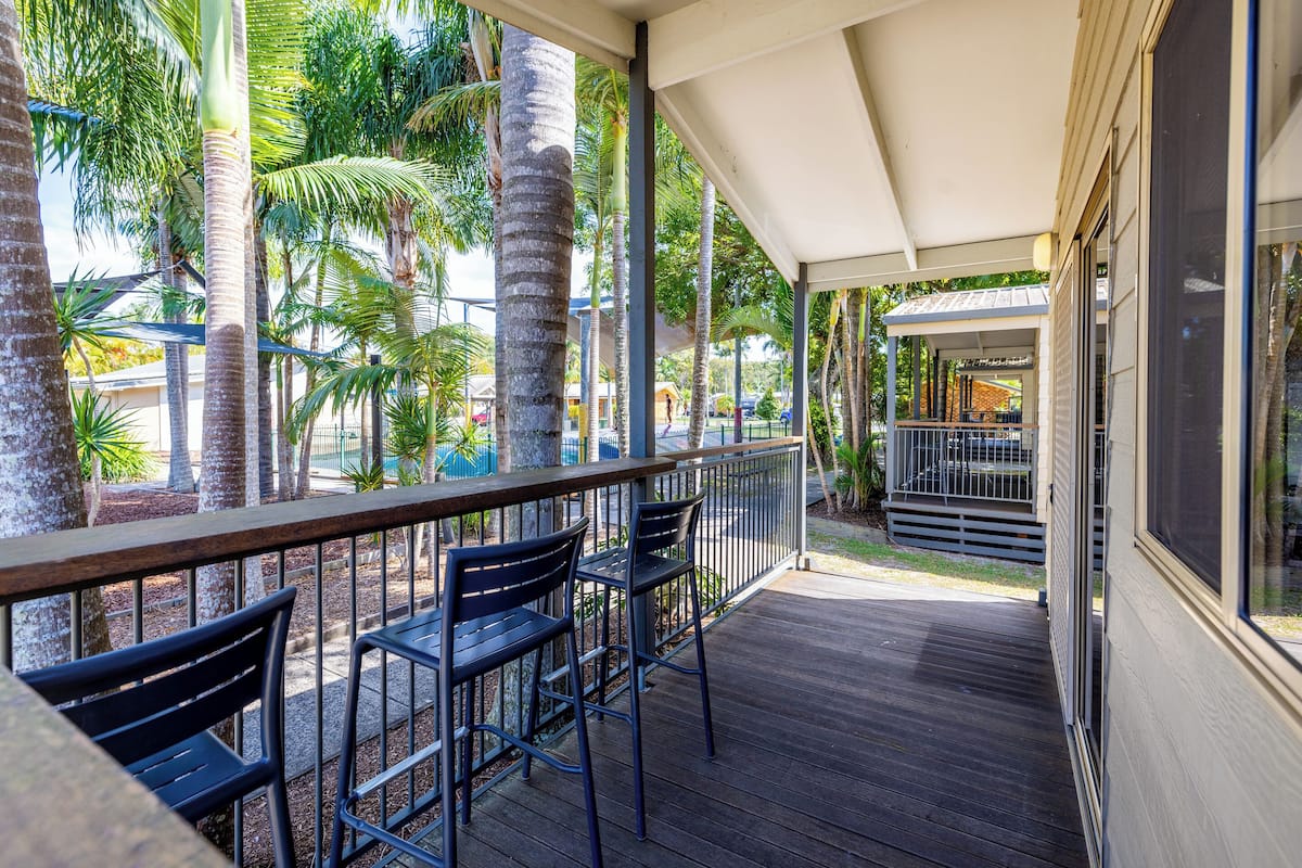A covered outdoor seating area with wooden tables and chairs, surrounded by palm trees and a view of a pool and buildings.