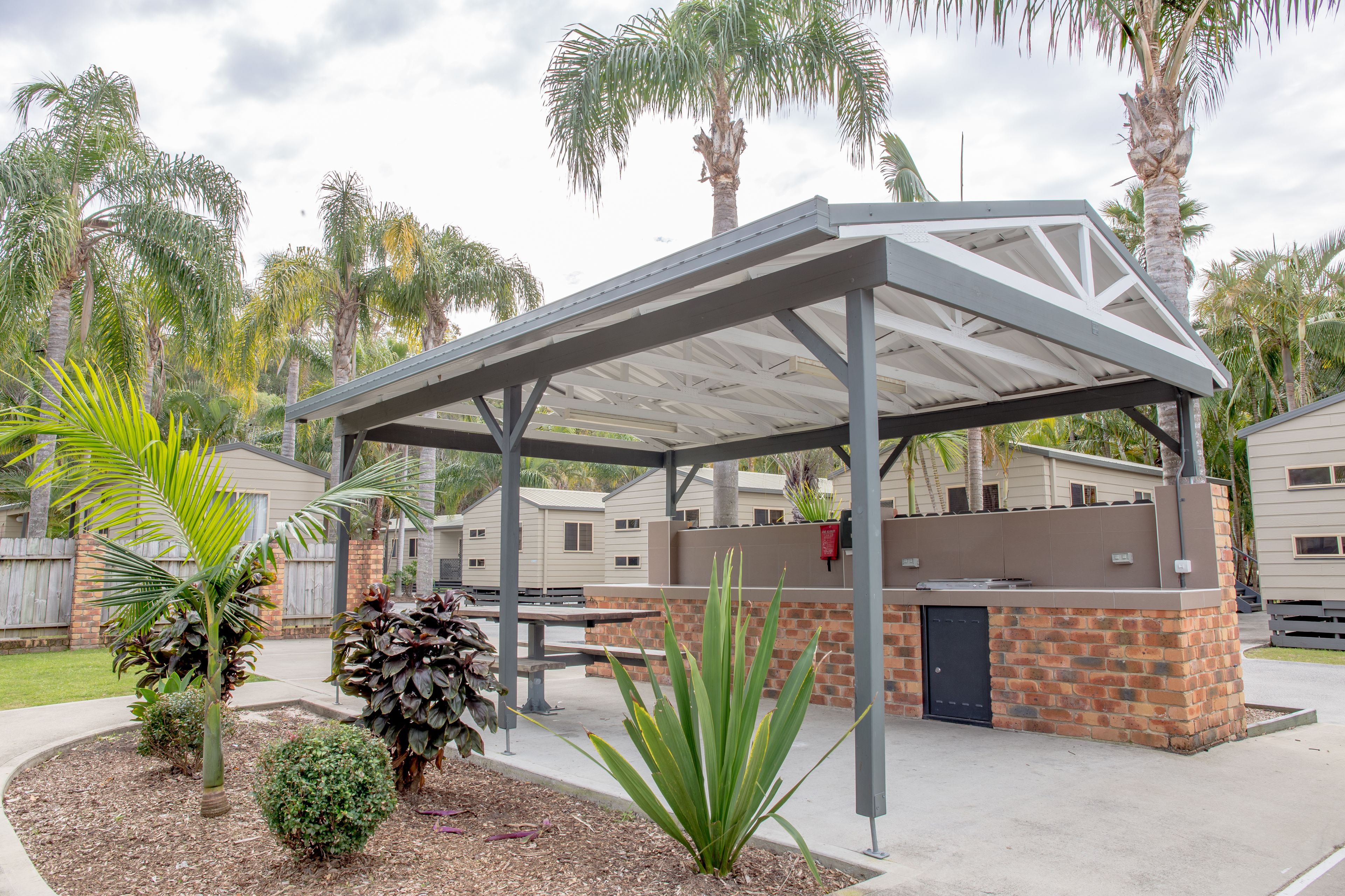 A covered outdoor patio area with a brick and metal structure, surrounded by palm trees and a residential setting.