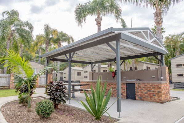 Un patio techado al aire libre con estructura de ladrillo y metal, rodeado de palmeras y un entorno residencial.