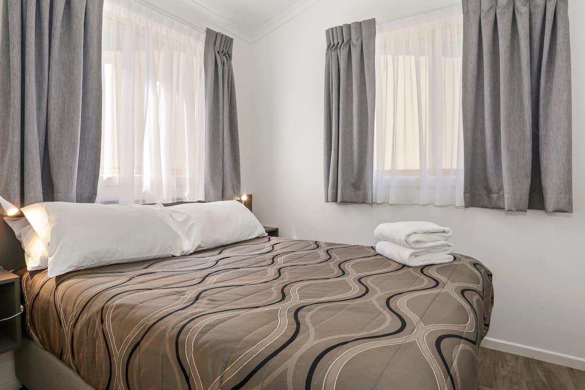 A hotel room with a bed, two windows with curtains, a bedside table, and a wall-mounted lamp.