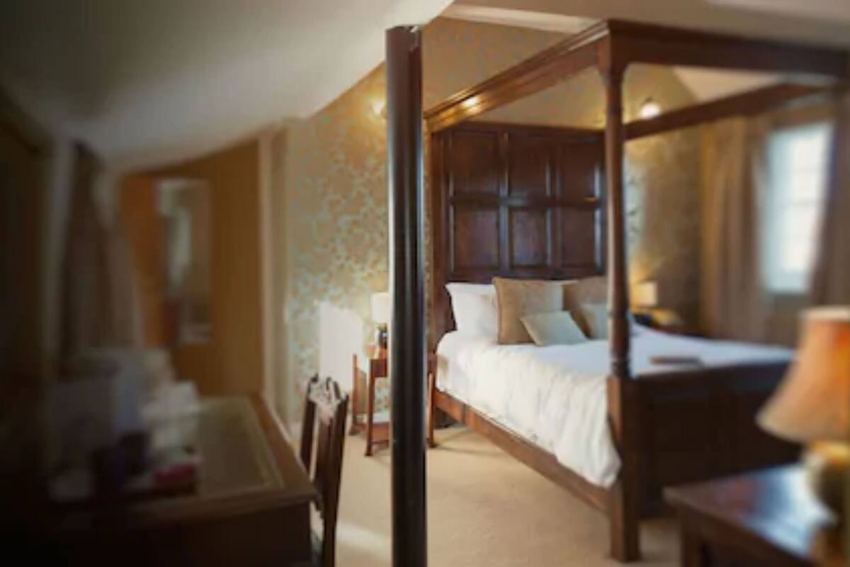 A four-poster bed with a wooden headboard, a bedside table, and a chair in a hotel room.