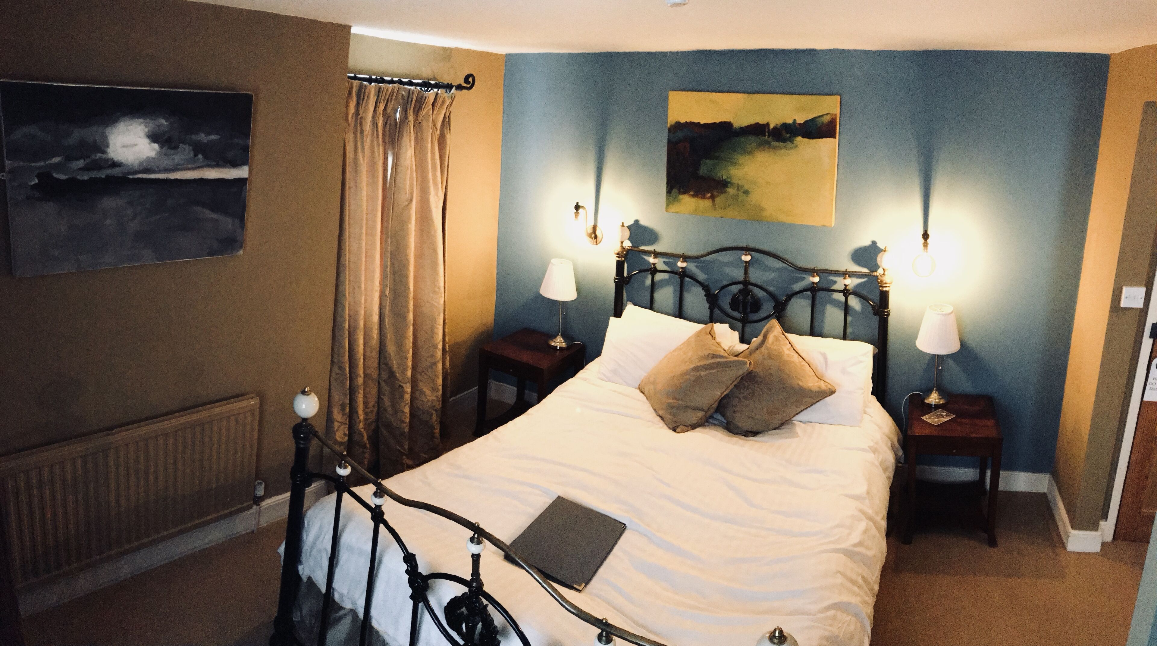 A neatly made bed with a pillow, a bedside table with a lamp, a painting on the wall, and a window with curtains.