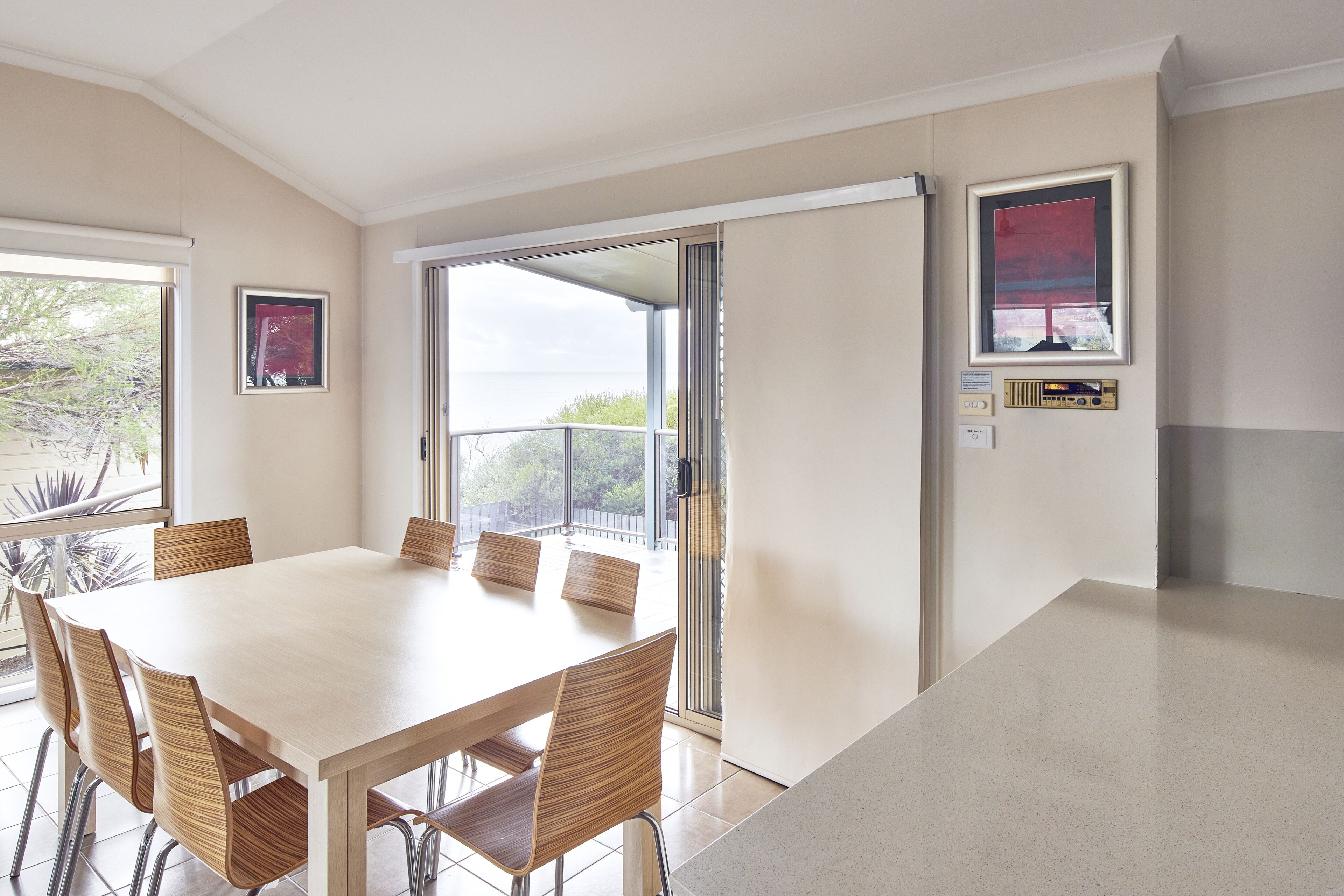 A dining area with a large table and chairs, a sliding glass door leading to a balcony, and framed pictures on the wall.