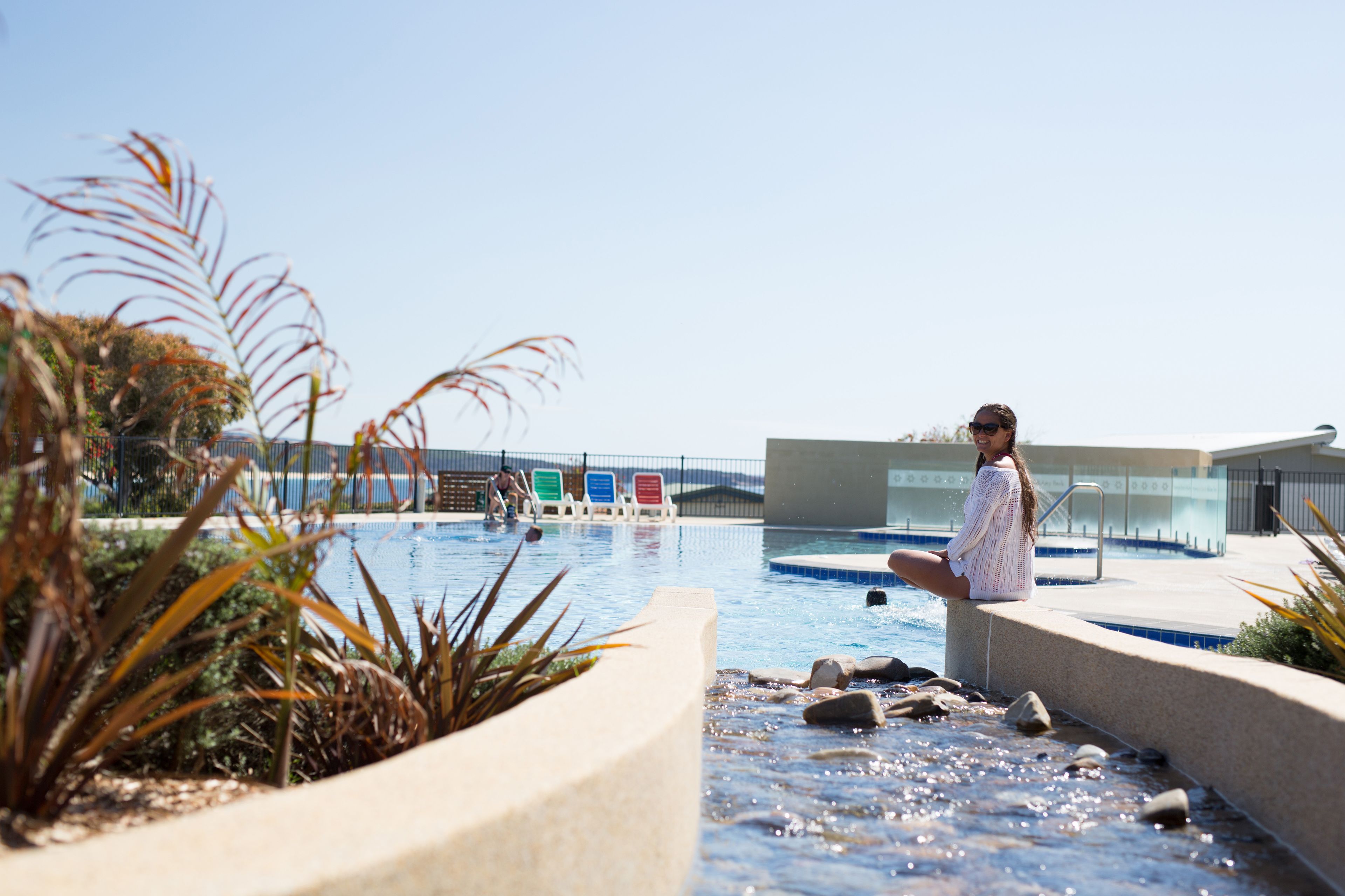 A pool area with a person sitting on the edge, surrounded by plants and a clear sky.