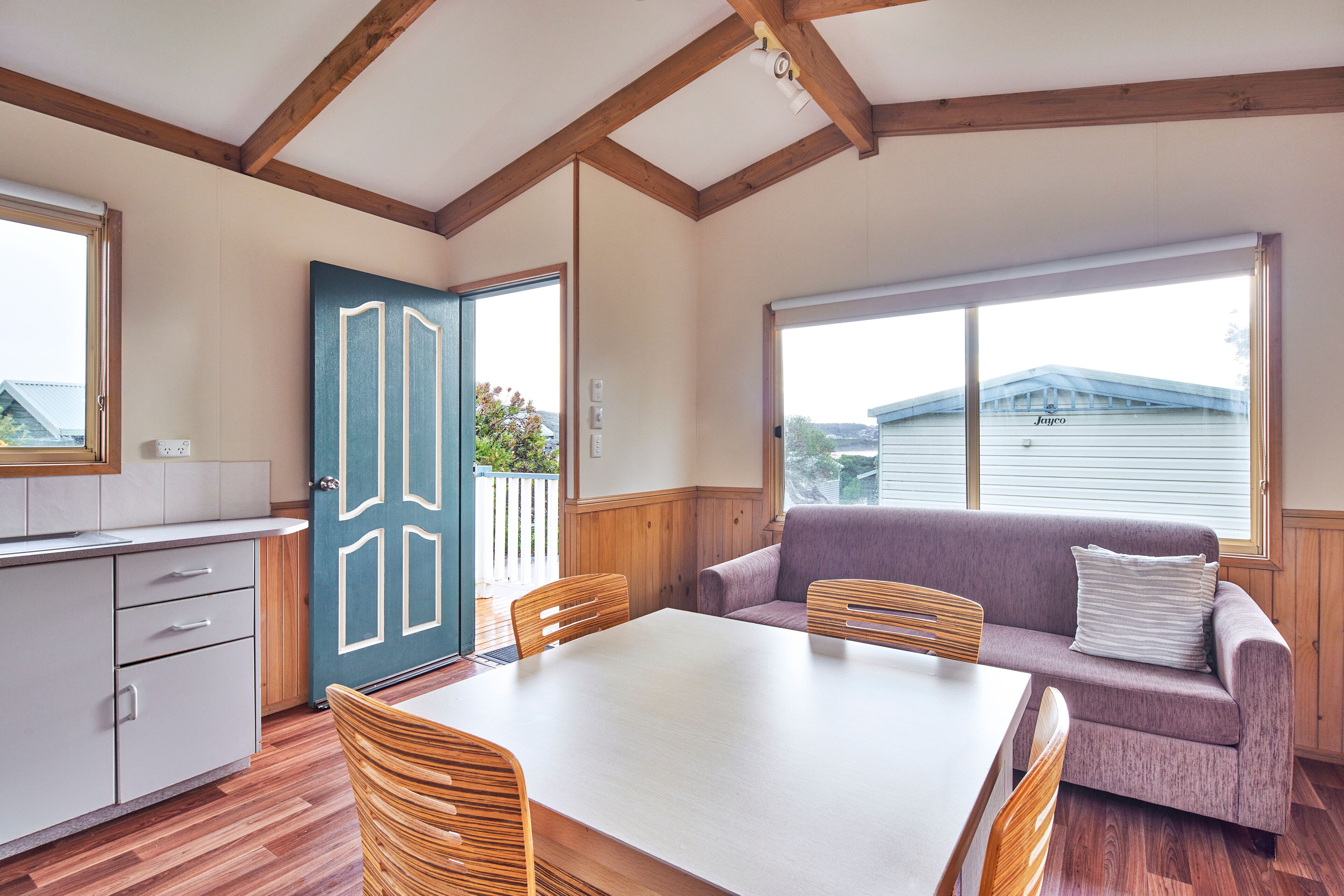 Una habitación con una mesa de comedor de madera, sillas, un sofá y una zona de cocina con armarios y una puerta.