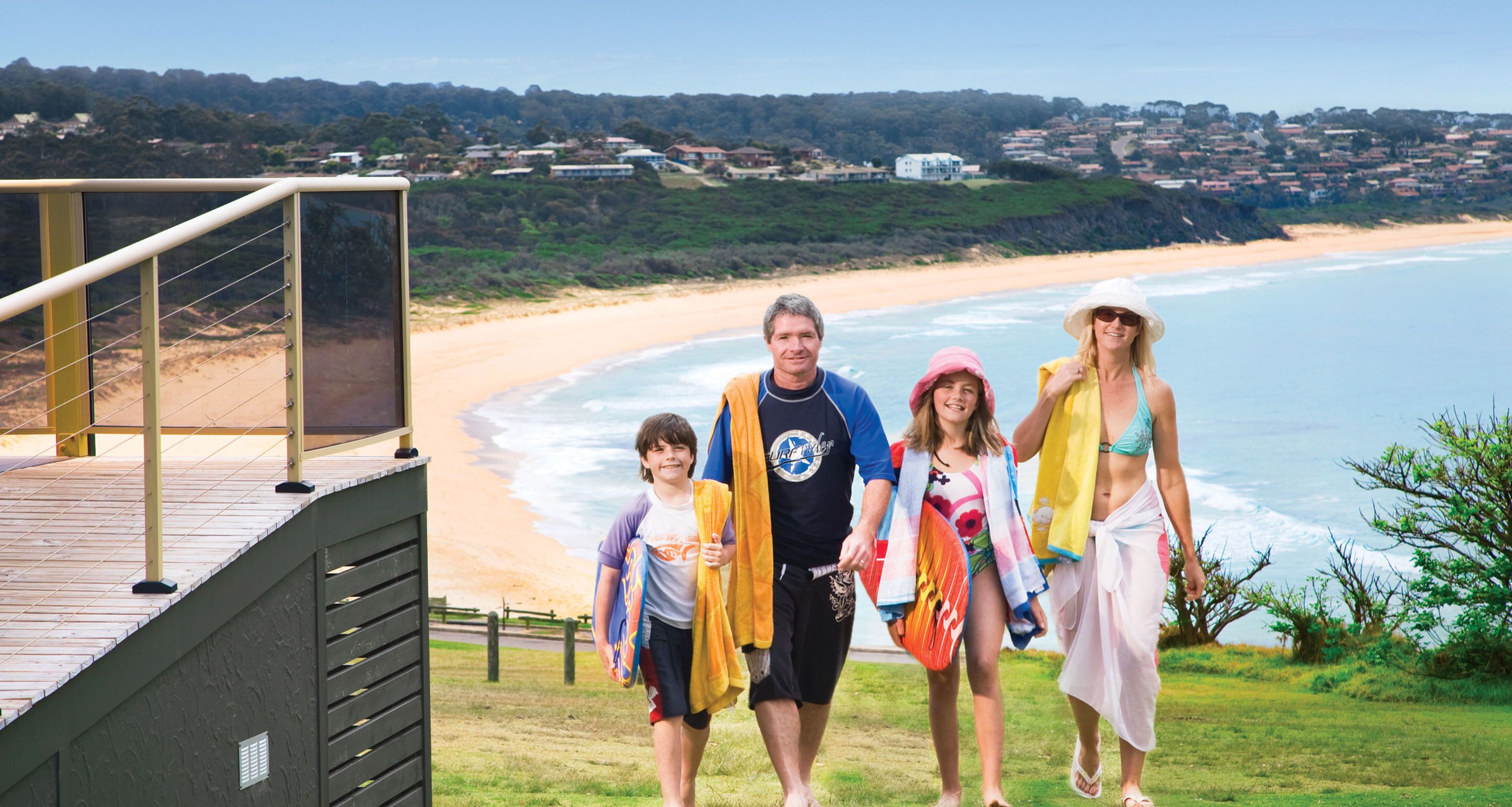 Una familia caminando sobre una terraza de madera con vistas a una playa, con casas al fondo.