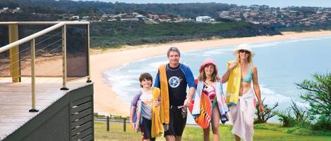 Una familia caminando sobre una terraza de madera con vistas a una playa, con casas al fondo.
