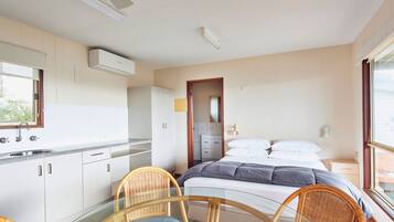 A compact hotel room with a kitchenette, a bed with white linens, and a dining area with wicker chairs and a glass table.