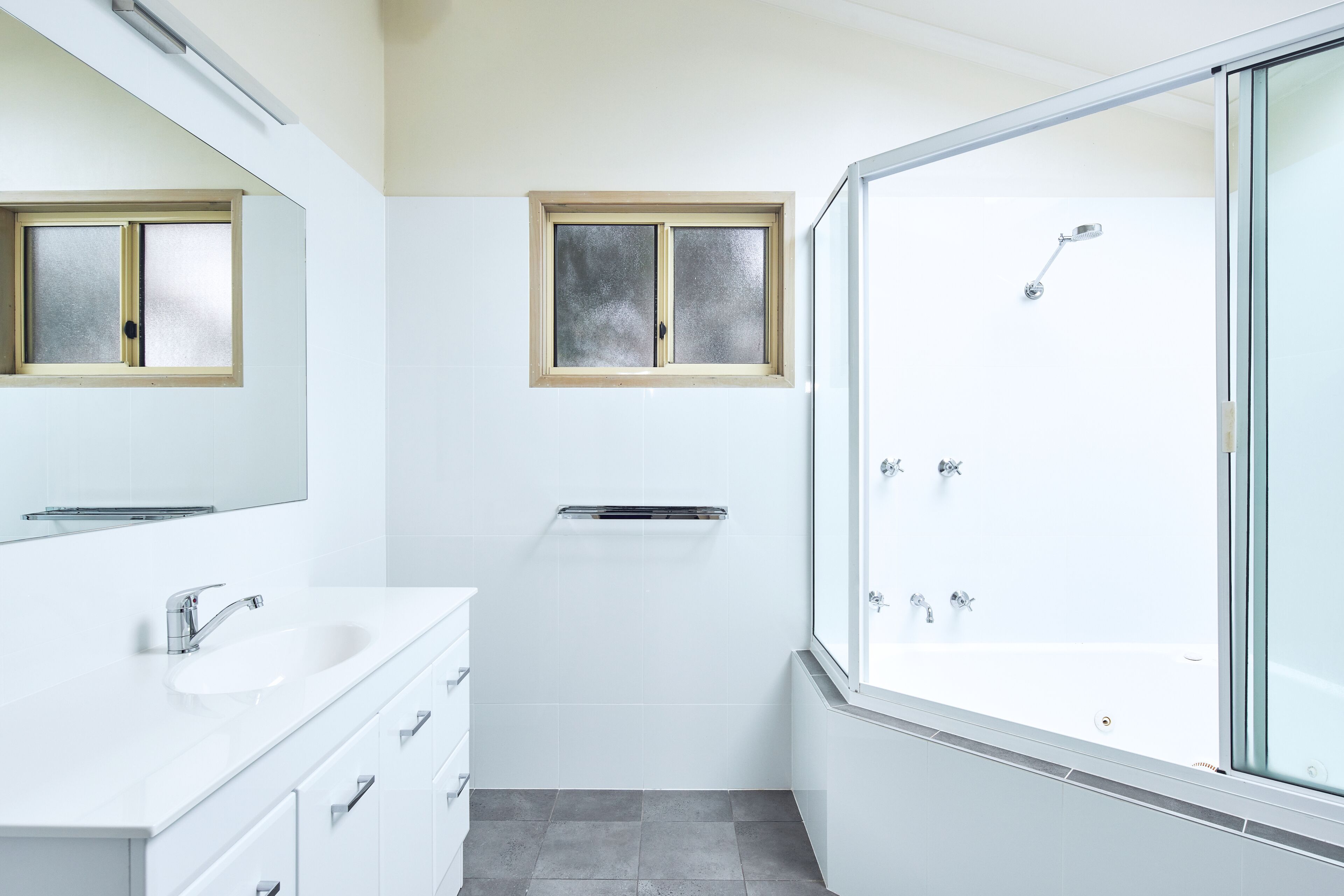 A modern bathroom with a white vanity, a large mirror, a window, and a glass-enclosed shower area.