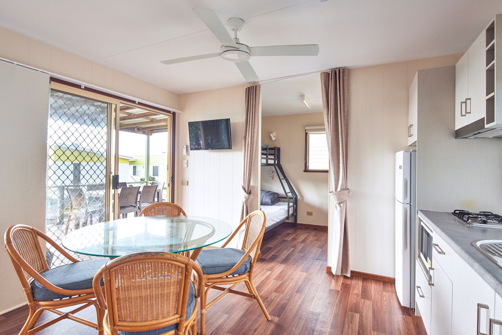 A compact room with a glass dining table, wicker chairs, a kitchenette, a ceiling fan, and a TV on the wall.