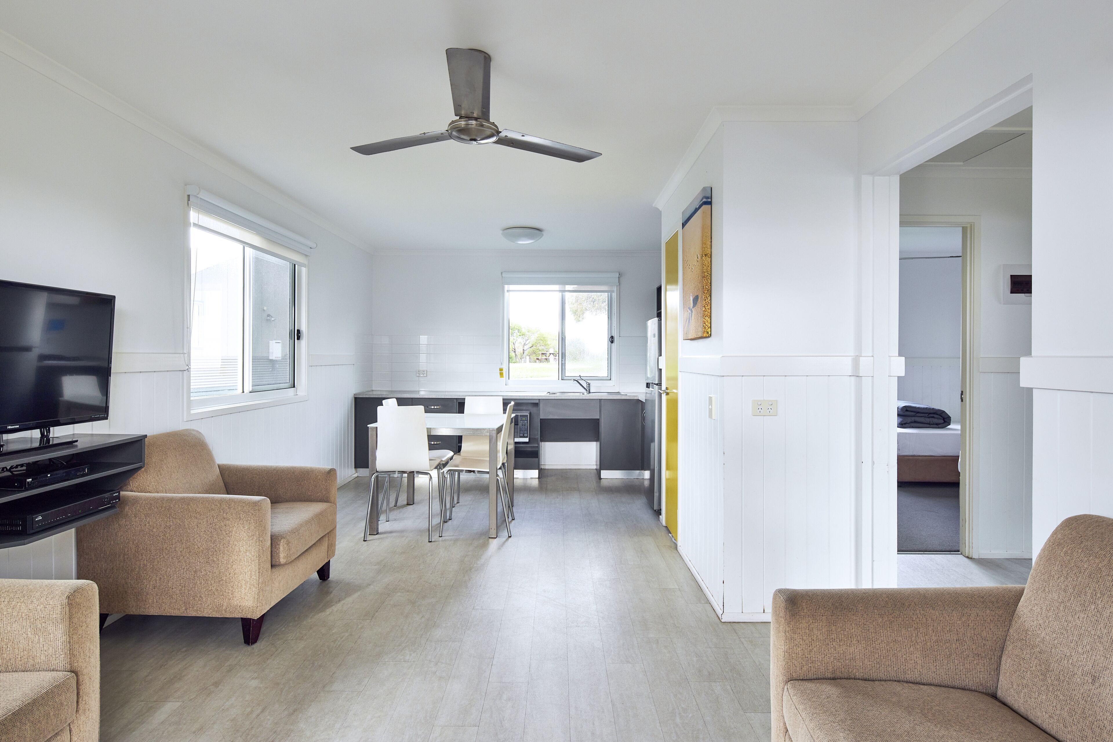 A modern living room with a flat-screen TV, beige sofas, a dining area with a table and chairs, and a kitchen in the background.
