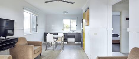 A modern living room with a flat-screen TV, beige sofas, a dining area with a table and chairs, and a kitchen in the background.
