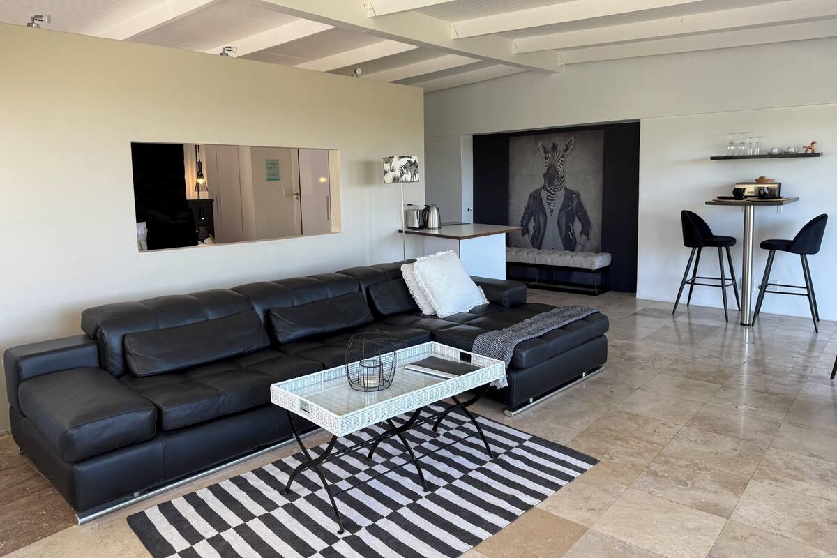 A modern living room with a black leather sofa, a white coffee table, a patterned rug, and a kitchen area with a zebra-themed artwork.